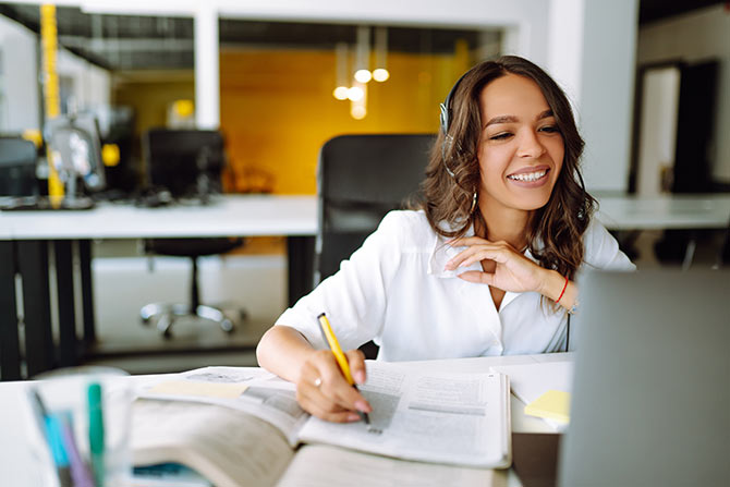 Bank Webinars; Smiling woman in white blouse, wearing headphones, works at a desk with open books and papers, holding a pencil. Bright and modern office setting.