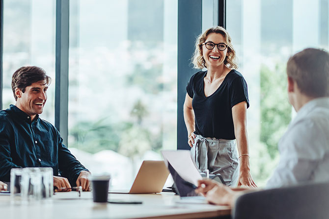 Inviting Questions to Spark Better Conversations; A cheerful woman stands, smiling and engaged, in a bright conference room with large windows. Two seated colleagues listen attentively, creating a collaborative atmosphere.